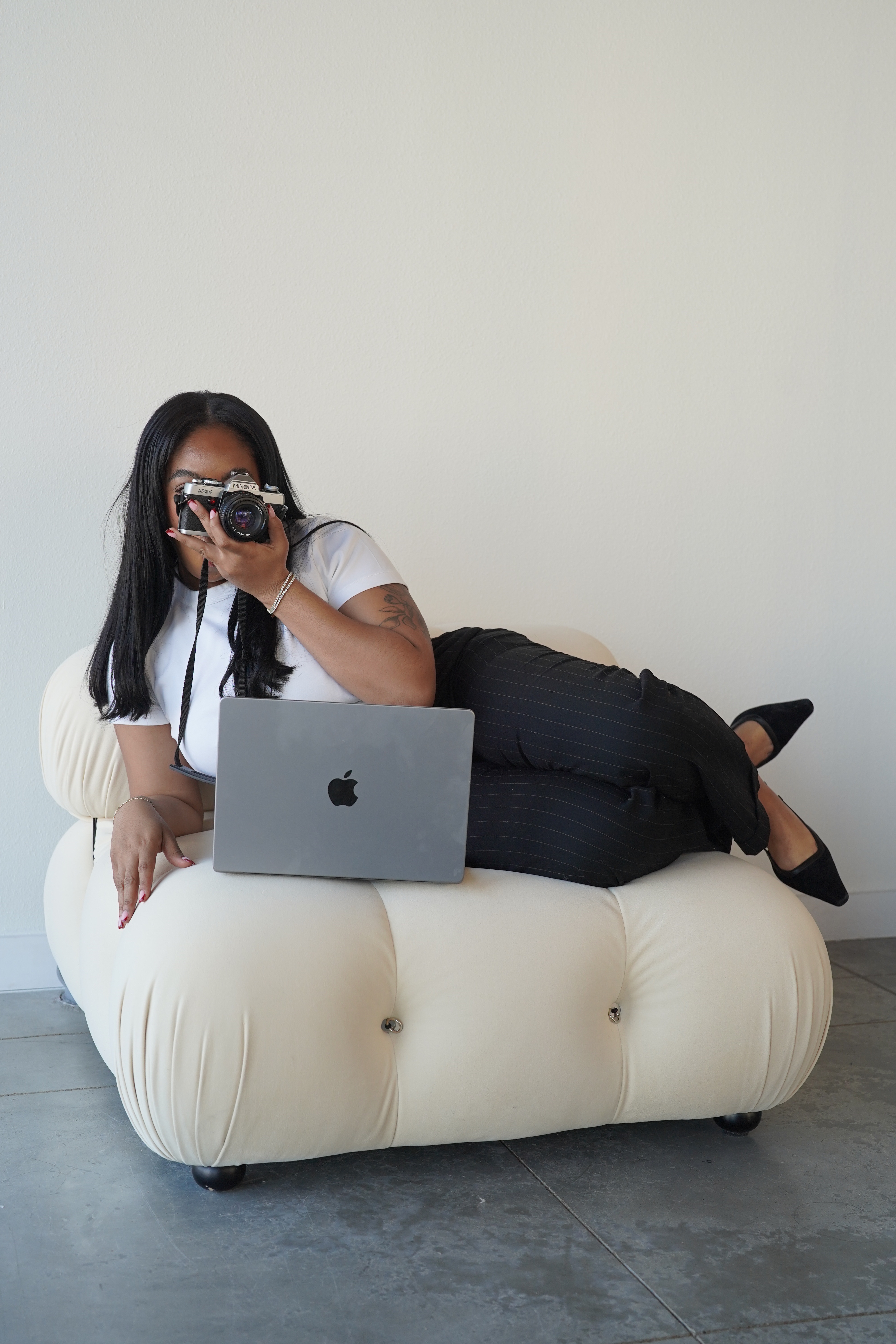 A woman reclining on a cream tufted sofa holding a vintage film camera with a laptop beside her