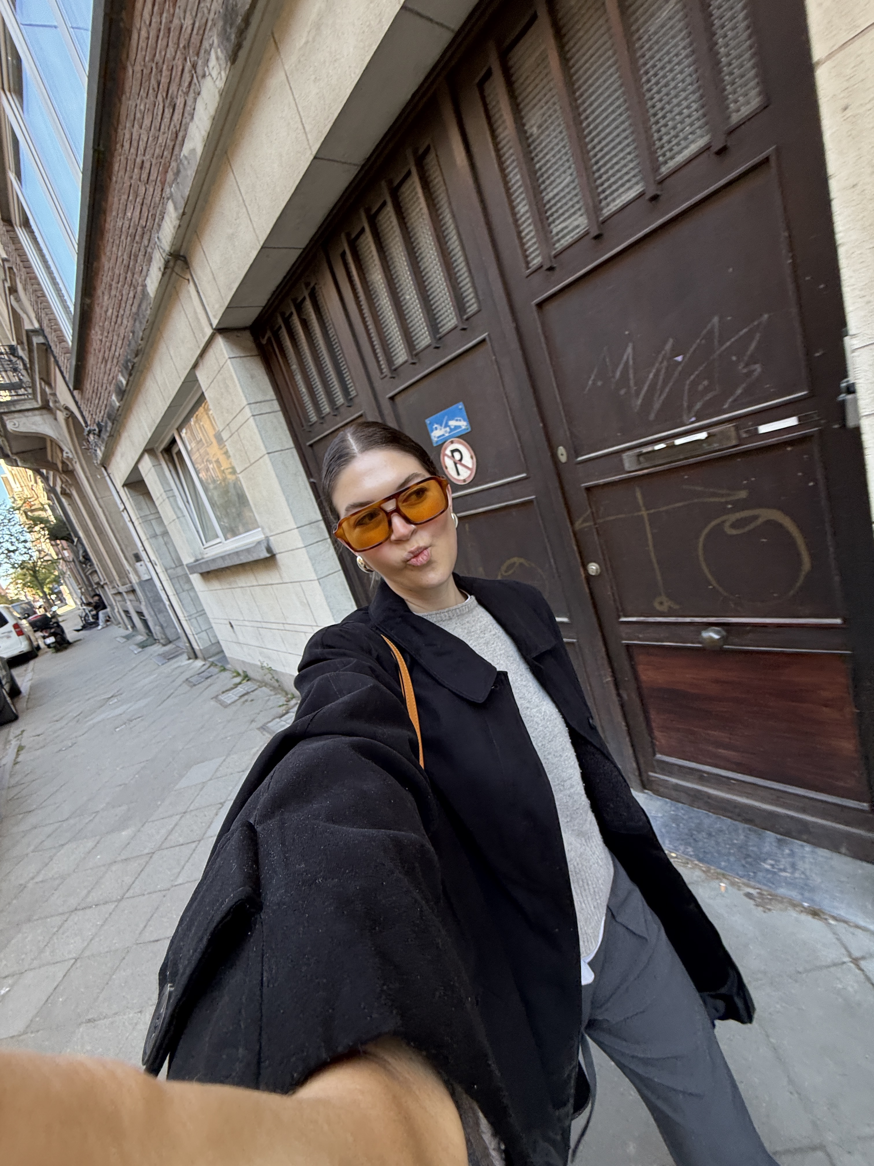 A woman in orange-tinted sunglasses taking a street-style selfie on an urban sidewalk